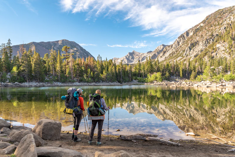 two backpackers stand in front of a calm alpine lake and mountains