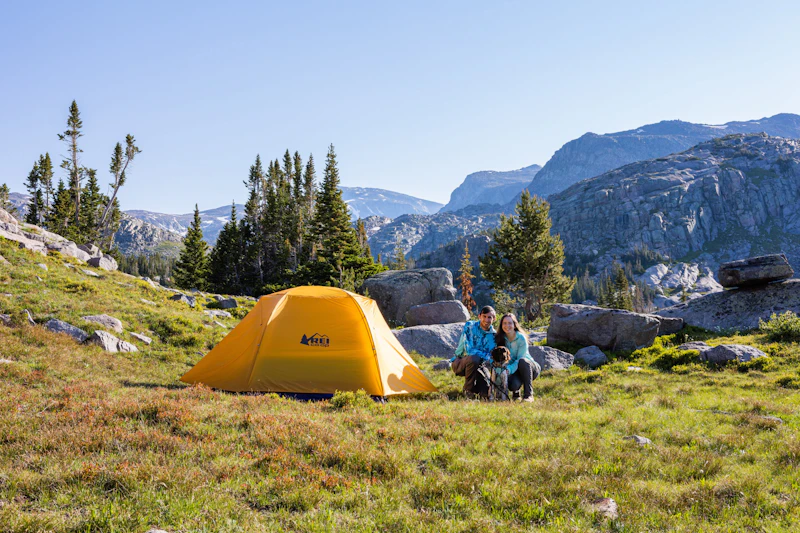 a man and woman pose next to their tent in a high alpine setting