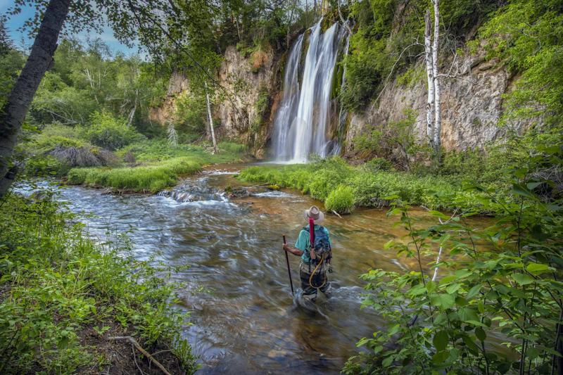 a fly fisherman stands in a pool beneath a forest waterfall