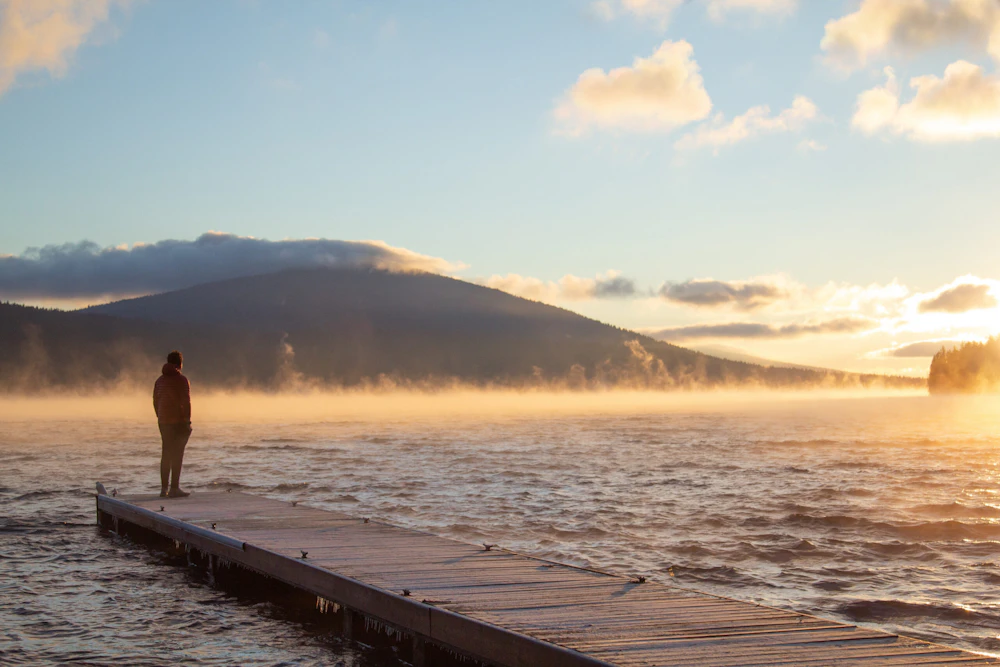 a person stands at the end of a dock at a lake a sunset