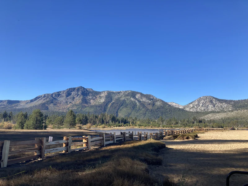 A new wooden wetland protection fence runs along the shoreline at Kiva Beach near Taylor Creek Visitor Center, with sweeping views of the Lake Tahoe Basin mountains under a clear blue sky.