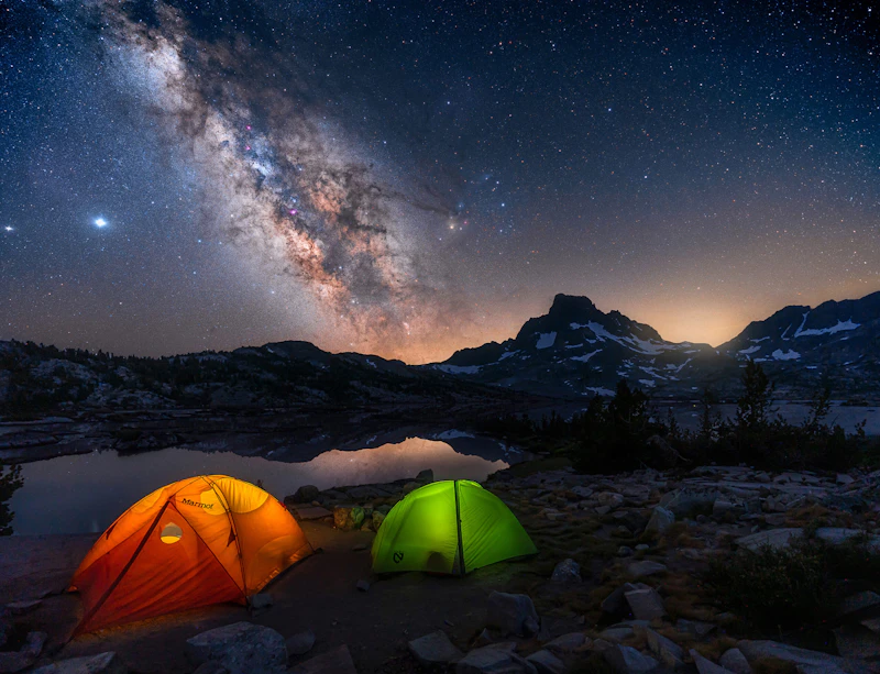 an orange and green tent illuminated from within beneath the milky way night sky and a large mountain