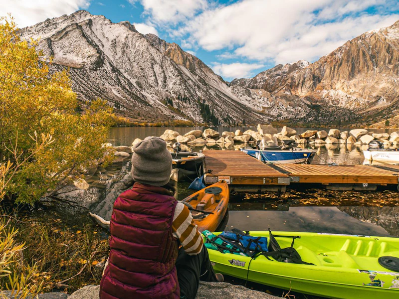 a woman sits in front of kayaks and a dock at a large alpine lake and mountains