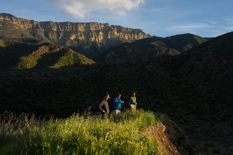 Three hikers stand on a grass cliff admiring the view.