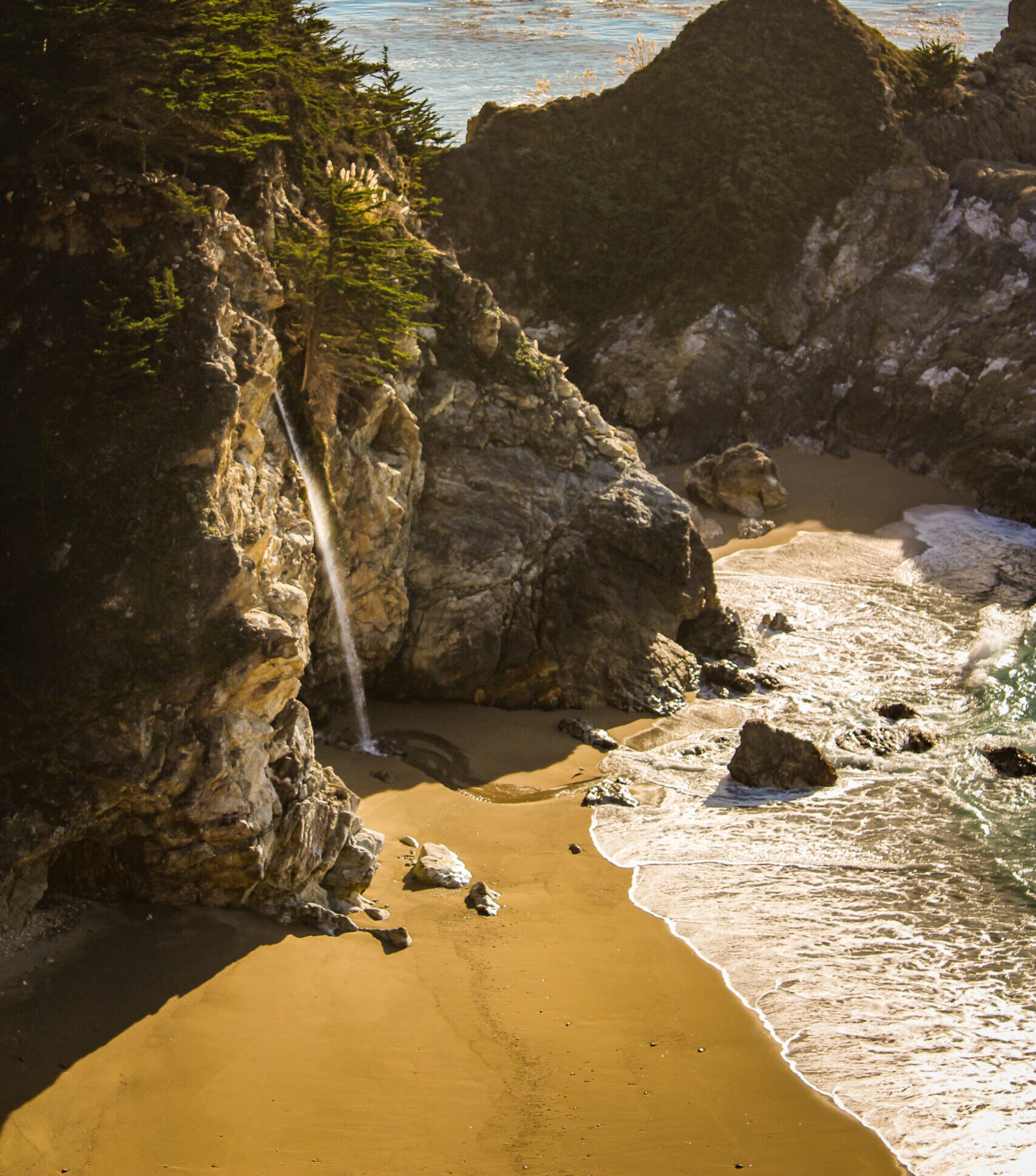 Waterfall from a cliff side flows into the ocean on a sandy beach.