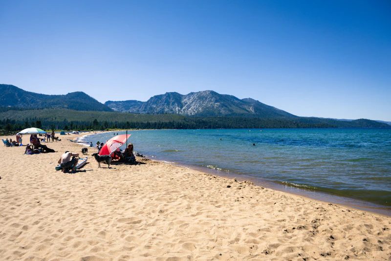 Visitors relax on a sandy beach along the shores of Lake Tahoe, with clear blue water and forested mountain peaks stretching across the horizon.