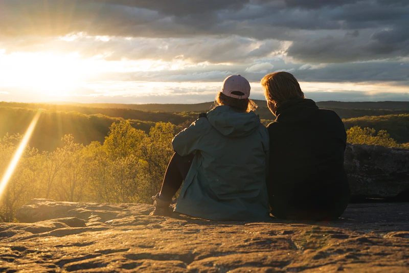 Couple sit together on a cliff at sunset.