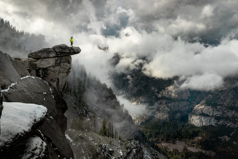 a hiker stands on a rocky precipice over a valley in foggy conditions