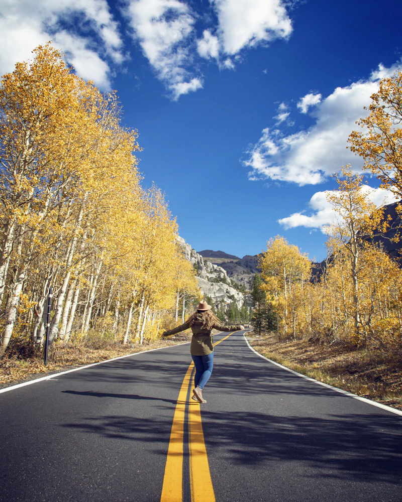 a woman with arms outstretched stands on a paved road in the mountains in fall