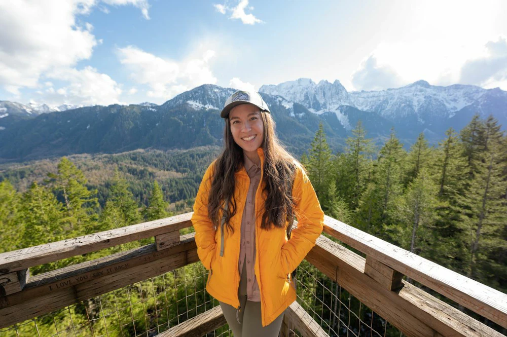 a woman in an orange jacket in an overlook in front of the forest and mountains