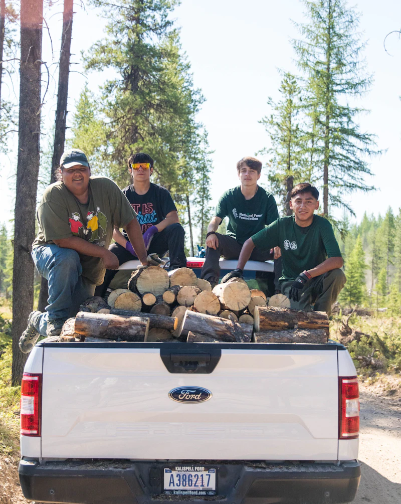 Members of the 2023 Mission Mountains Youth Crew sit on the back of a pick up truck with firewood removed from the forest.