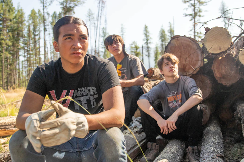 three young men sit on a log pile in the forest