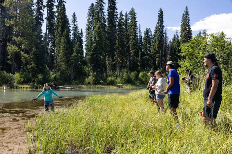 Young adults stand at the edge of a marshy lake listening to a woman