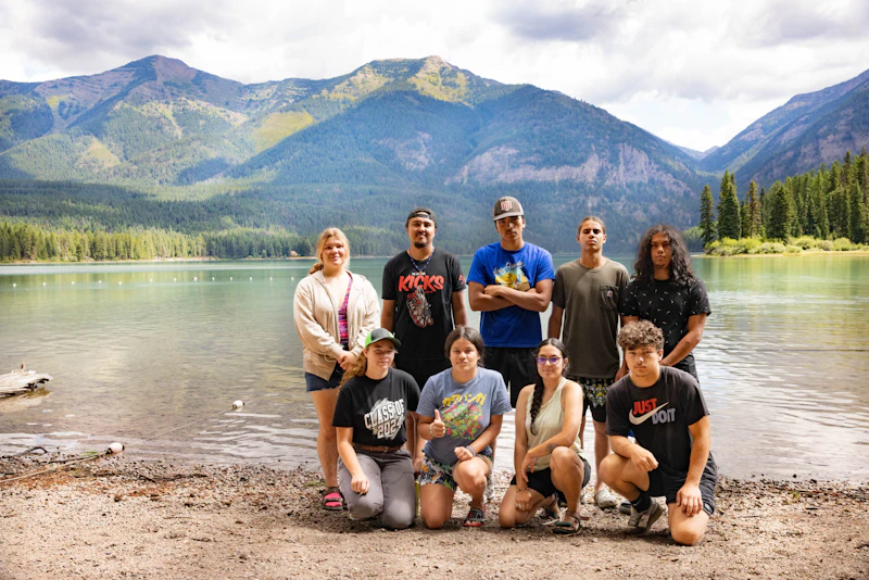a group of young adults pose as a group in front of a lake and mountains
