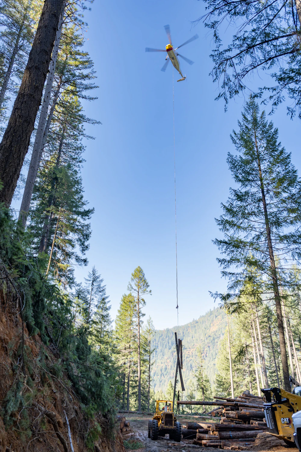 Helicopter lowers its cable to crew members to secure logs for air travel.