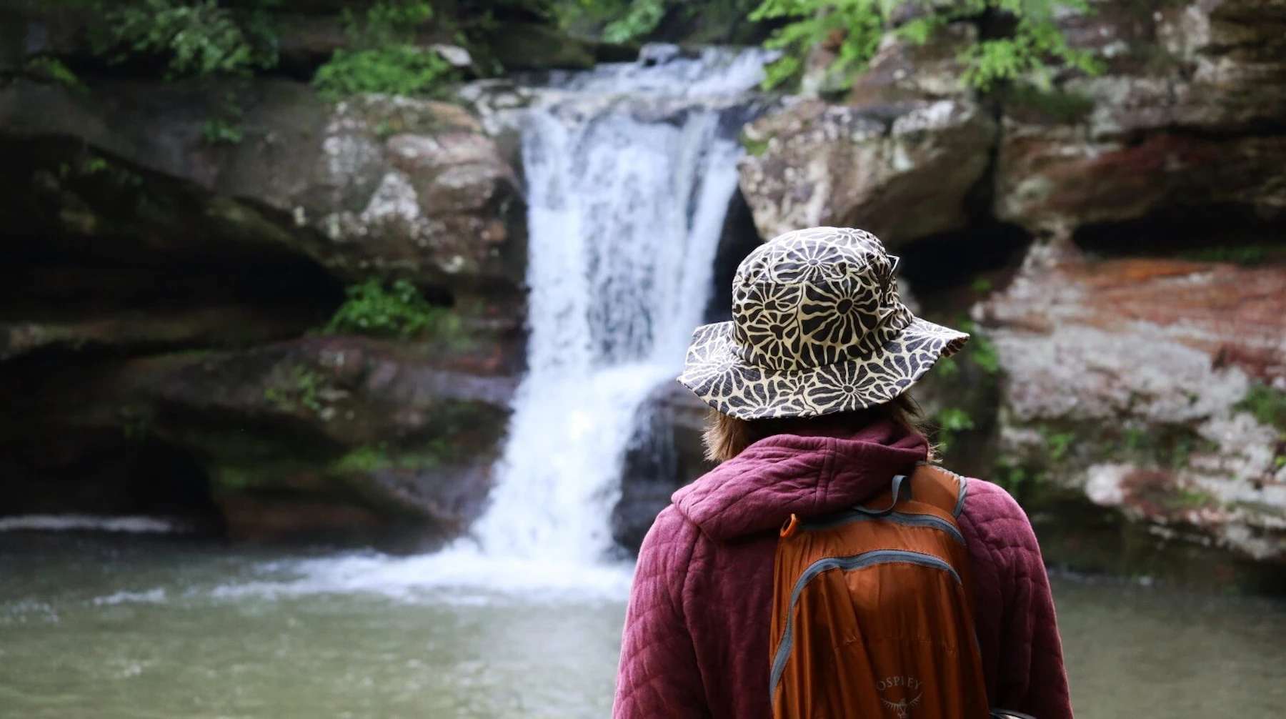 A hiker in a wide-brimmed sun hat pauses to watch a waterfall tumble over moss-covered rock in Wayne National Forest.